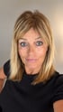 Porträt einer Frau mit blonden, schulterlangen Haaren, blauen Augen und schwarzem T-Shirt vor grauem Hintergrund.