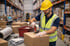 Arbeiter in gelbem Helm und Warnweste klebt ein Paket in einem Lager.