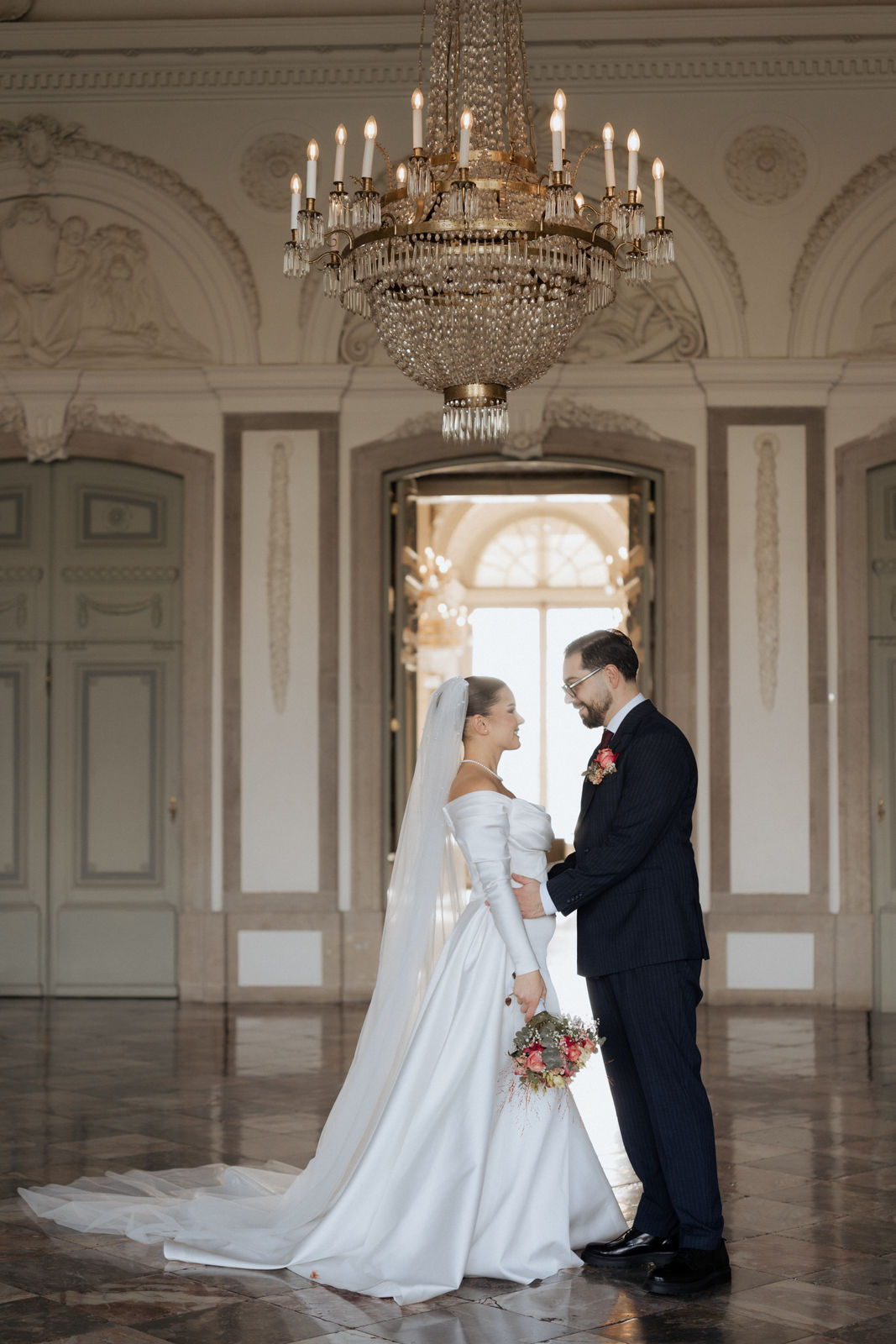 Brautpaar steht sich unter dem Kronleuchter im Schloss Benrath in Düsseldorf gegenüber.