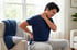 A man sits in an armchair clutching his neck and lower back with a grimacing expression of pain.