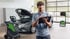 Ein Techniker mit Tablet steht in einer Werkstatt vor einem Auto mit geöffneter Motorhaube und Diagnosegeräten.