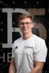 Lächelnder Physiotherapeut mit Brille und weißem Adidas-Shirt mit dem Logo „THERAPIER this is PHYSIO“ und Pier5 