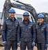 Drei lächelnde Bauarbeiter in Bausantec-Arbeitskleidung mit blauen Helmen stehen vor einem Bagger der Marke Bausantec.