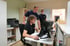 Drei lächelnde Männer arbeiten gemeinsam an einem Computerbildschirm in einem hellen Büro mit Schreibtisch und Drucker.