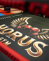 Nahaufnahme eines Boxrings mit schwarzem Boden, goldener "HORUS GYM"-Aufschrift und Logo aus Boxhandschuhen mit Flügeln.