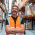 Lächelnder Lagerarbeiter mit Bart in orangefarbener Warnweste und schwarzem Hemd, Arme verschränkt.