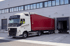Ein weißer Volvo-LKW mit rotem Anhänger fährt rückwärts an eine Laderampe. Fahrer schaut aus dem Fenster.