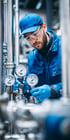 Ein Mann in blauer Arbeitskleidung, Mütze und Schutzbrille überprüft Manometer an Industrieanlagen.