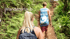 Wanderurlaub: Eine blonde Frau mit Rucksack folgt einem Mann auf einem bewaldeten Wanderweg.