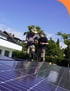 Handwerker installieren Solarmodule auf einem Ziegeldach, mit Gerüst und blauer Plane im Vordergrund.