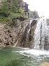 Eine Person in einem Neoprenanzug springt von einem Felsen in einen Wasserfall, zwei weitere Personen stehen oben.