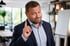 Mann im Anzug mit erhobenem Zeigefinger und fragendem Gesichtsausdruck in einem Büro.