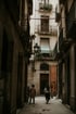 Two people walk down a narrow, old city alleyway with tall buildings, balconies, and a streetlamp in Spain.