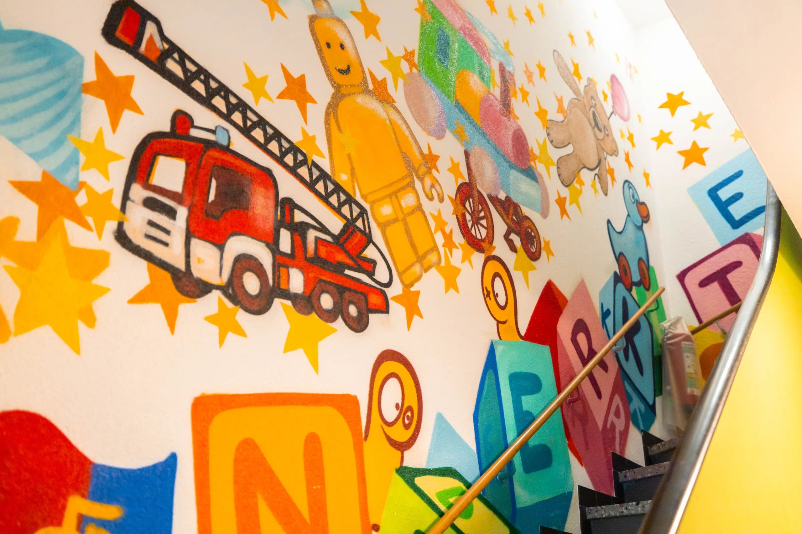 Bunte Graffitiwand im Treppenhaus der Kita mit Feuerwehrauto und Buchstabenwürfeln.