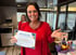 A smiling woman in a red cardigan holds a work anniversary certificate for Kara McGahey next to a bouquet of flowers.