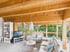 Modernes Wohnzimmer in einem Holzhaus mit sichtbaren Deckenbalken, hellen Sofas, Fernseher und großer Fensterfront zum Garten.