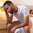 Man sitting on bed looking sad, woman blurred in background – depicting Erectile Dysfunction.