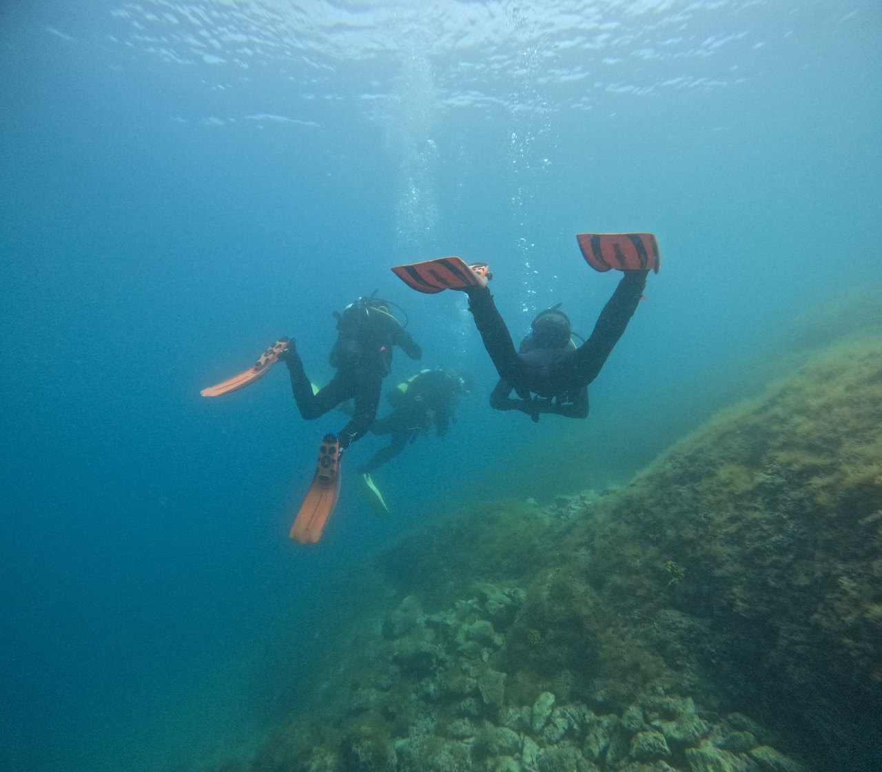Three divers descending along a rocky slope in turquoise water