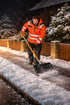 Ein Mann in orangefarbener Warnjacke schiebt nachts Schnee von einem Bürgersteig.