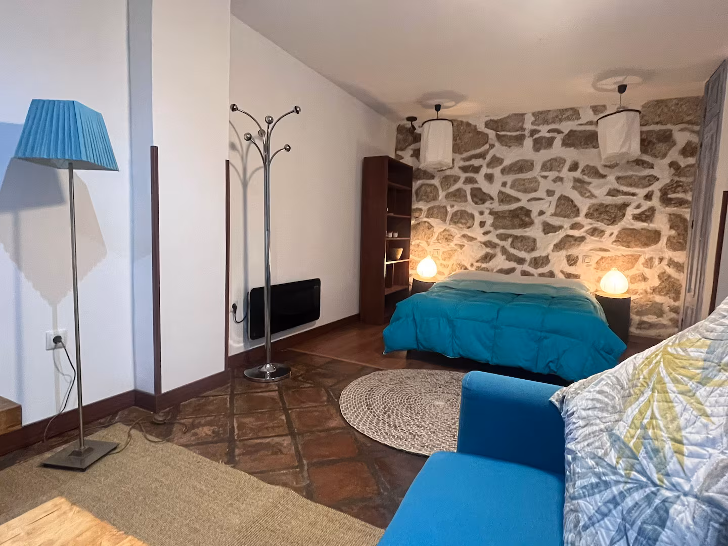 Habitación de hotel con cama azul, pared de piedra, sofá azul, lámpara de pie y perchero metálico.