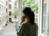 Frau im grauen Pullover telefoniert am Fenster mit Blick auf einen Innenhof.
