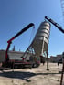 Zwei LKW-Kräne heben gemeinsam ein großes Metallsilo auf einem Industriehof an.