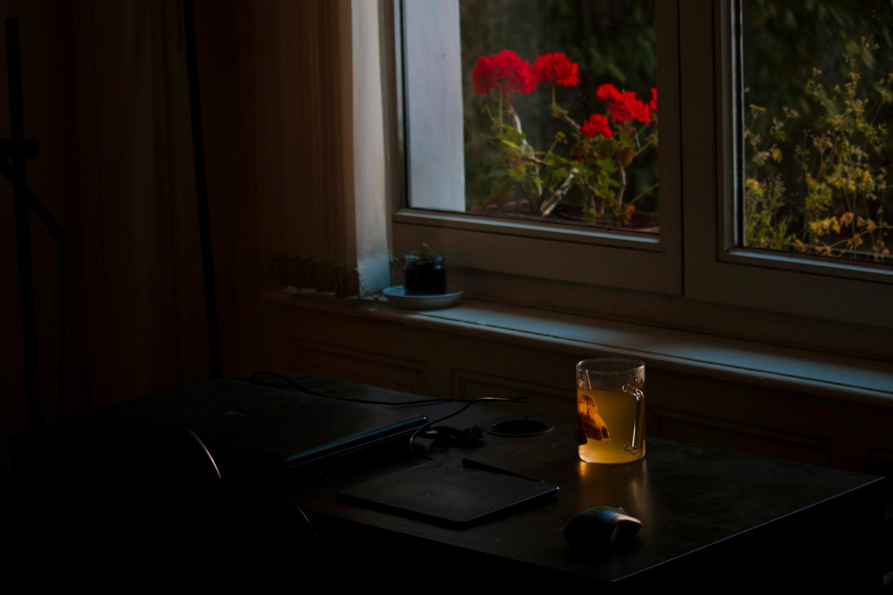 Dämmriger Raum mit einem Schreibtisch am Fenster, darauf ein Glas mit bernsteinfarbenem Getränk im letzten Abendlicht, draußen rote Geranien.