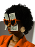 A person with dark curly hair wears heart-shaped sunglasses, one-dollar bills tucked under each lens, and an orange blazer.