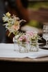 Three small glass vases with various colorful flowers sit on a white tablecloth with a wine glass.