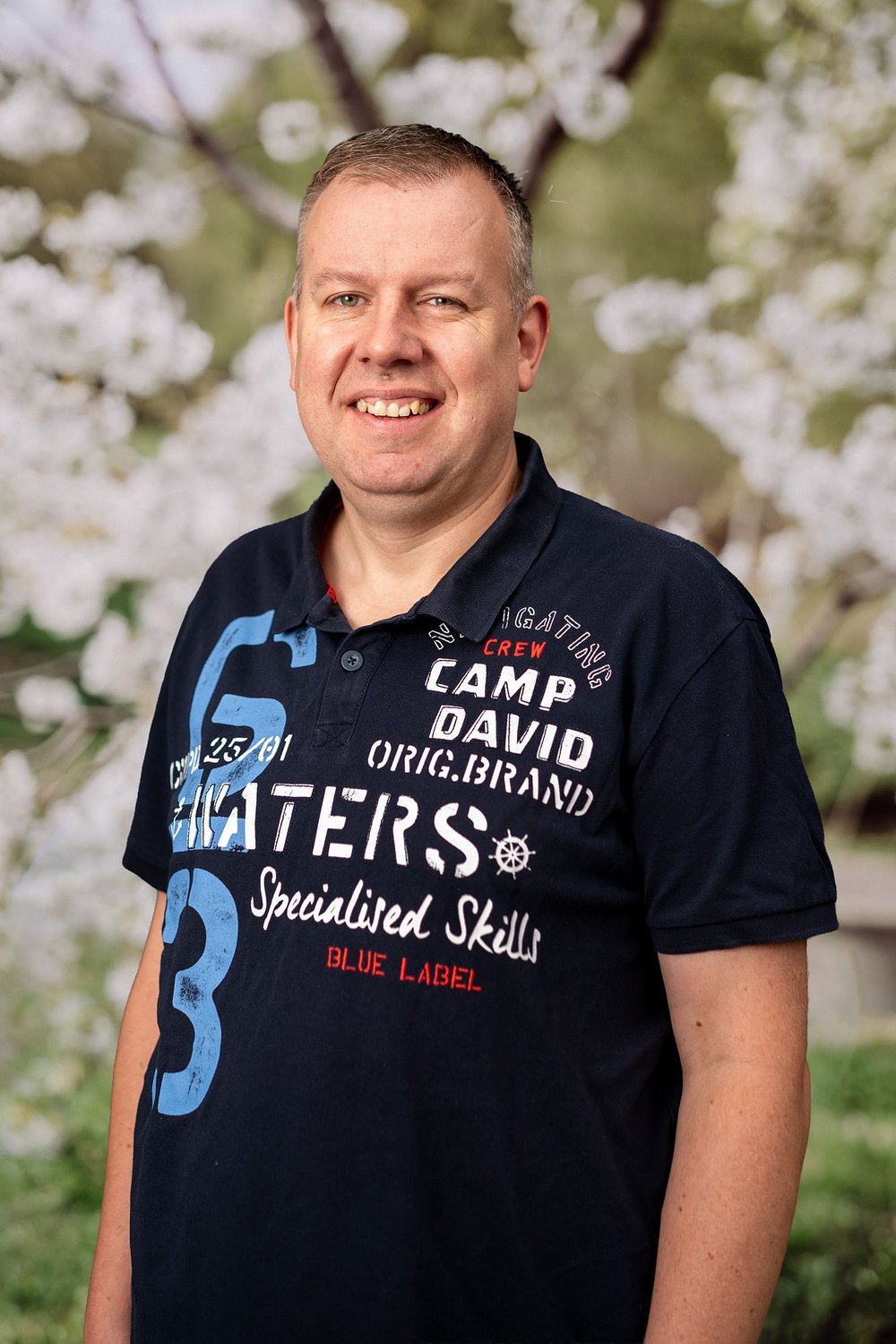 Mann mit Bart und Brille in rotem Pullover mit Reißverschluss und "GREEN LAND-PRO MOWING"-Logo vor weißen Blumen.