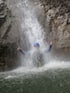Person in Neoprenanzug unter einem Wasserfall, der beide Daumen hochhält.