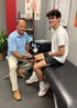 A physical therapist applies a device to a smiling young man's knee in a clinic room.