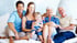 Drei Generationen einer Familie sitzen lächelnd zusammen auf einem hellen Sofa.