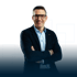 Mann mit Brille in blauem Hemd und dunklem Sakko lächelt mit verschränkten Armen.