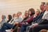 Junge Frau im grauen Blazer sitzt bei einer Konferenz, hält Papiere und blickt nach vorne zur Rednerin.