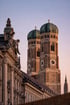 Zwei braune Kirchtürme mit grünen Kuppeln und Zifferblättern ragen über einem verzierten Gebäude und einem rosafarbenen Himmel empor.