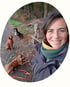 Lächelnde Frau macht ein Selfie im Wald mit vier angeleinten Hunden, die im Hintergrund auf einem Waldweg sitzen.