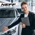 Ein lächelnder Verkäufer mit Brille und schwarzem Anzug präsentiert ein silbernes Auto; NEFF Autohaus NEFF GmbH Logo.