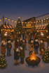 Menschen auf einem Weihnachtsmarkt mit Lichterketten, Tannenbaum, Marktständen und einer Feuerschale in der Dämmerung.