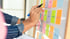Eine Person schreibt mit einem Marker auf bunte Haftnotizen an einem Whiteboard während eines Brainstormings.
