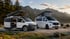 Zwei weiße Campervans mit aufgestelltem Dachzelt, einer mit offener Schiebetür, vor Bergen und See.