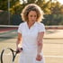 A woman in white tennis apparel looks down at a large purple and red bruise on her forearm while holding a tennis racket.