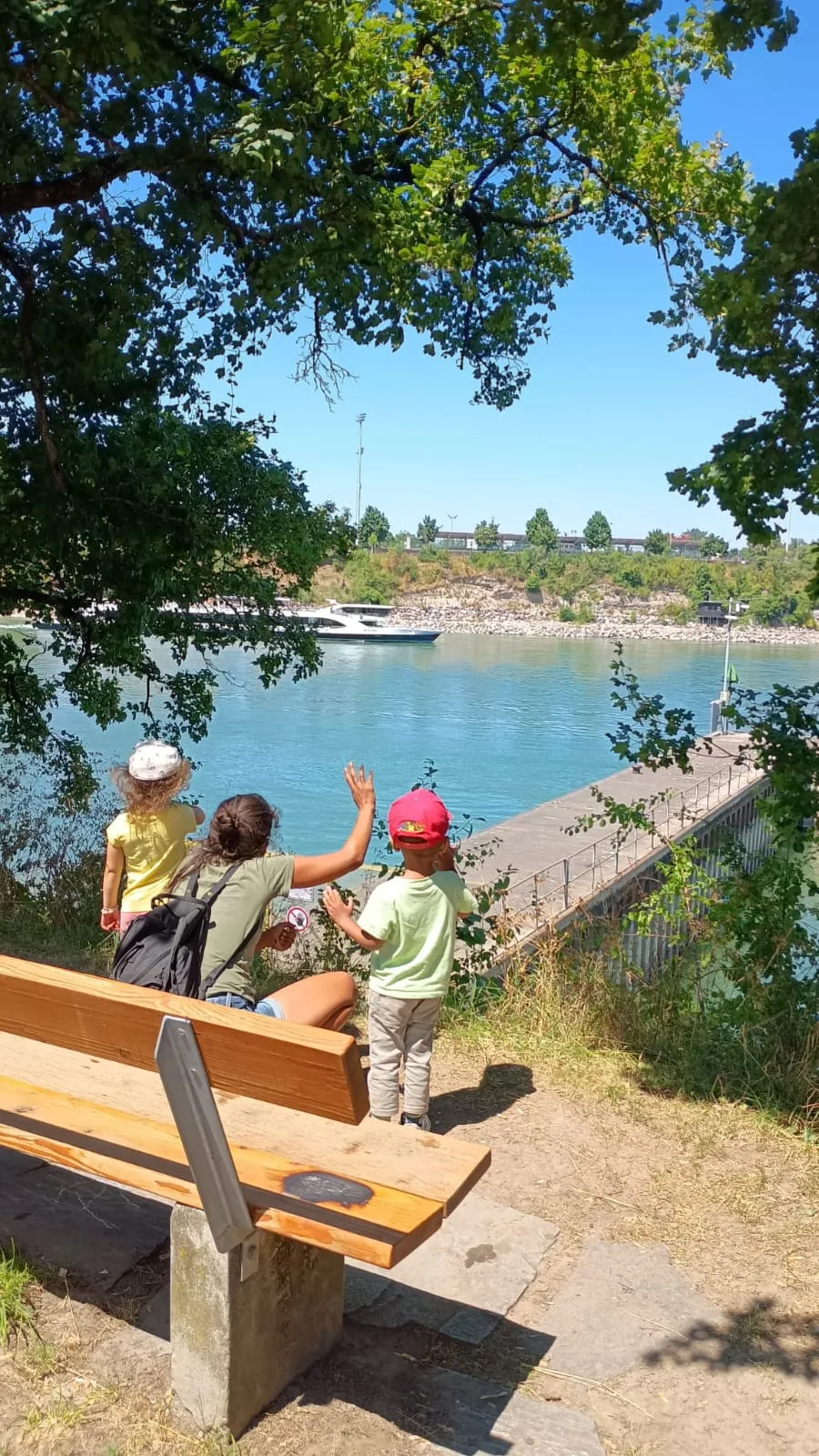 Kinder sitzen auf einer Bank am Rhein und winken einem Schiff.