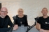 Drei Podcaster, zwei Männer und eine Frau, sitzen vor Mikrophonen an einem Tisch.