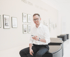 Ein lächelnder Mann mit Brille und Smartphone sitzt in einer Galerie vor Fotografien der Fred Stein Ausstellung.