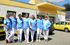Sieben lächelnde Frauen in blauen Uniformen stehen vor einem gelb-weißen Gebäude und zwei gelben Autos.