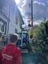 Ein Kran hebt einen Baum über ein Wohnhaus, während ein Mitarbeiter von GartenGlück und ein Minibagger die Arbeit begleiten.