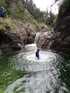 Person im Neoprenanzug mit Helm im Wasser, umgeben von Felsen und einem Wasserfall.