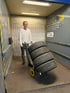 Ein lächelnder junger Mann schiebt eine Sackkarre mit vier gestapelten Autoreifen in einem Korridor.