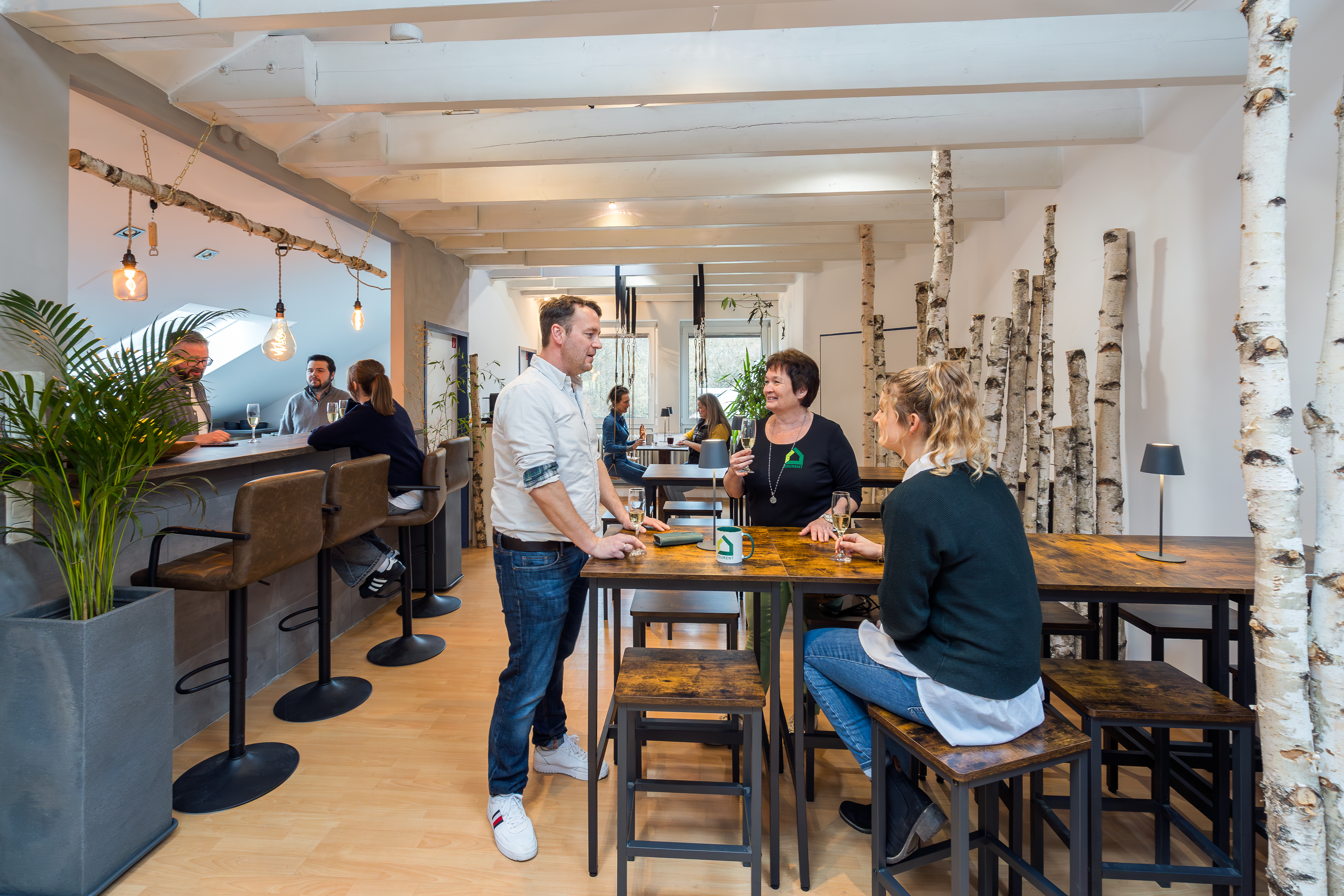 Menschen unterhalten sich in einem hellen Raum mit hohen Tischen, Barhockern, Birkenstämmen und einer Bar.